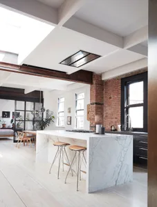 A modern kitchen featuring a white marble island, glass pendant lights, and an exposed brick wall.