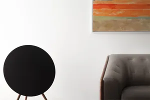 A minimalist living room featuring a black spherical speaker on a wooden tripod and a taupe-colored upholstered chair.