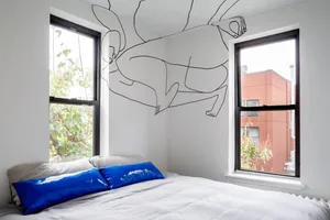 A minimalist bedroom with white walls, a bed with a blue pillow, and a wall mural of a black line drawing of a person