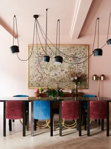 A modern dining room featuring a wooden table surrounded by red and blue velvet chairs, illuminated by black pendant lights a