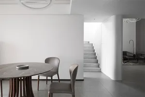 Minimalist dining room featuring a round marble table with gray chairs, concrete stairs, and a white wall.