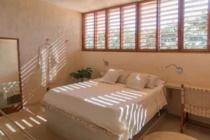 A minimalist bedroom featuring a concrete bed platform, white bedding, and closed shutters with sunlight filtering through.