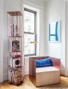 A minimalist living room featuring a metallic bookshelf, a comfortable chair with a blue cushion, and a framed window view.