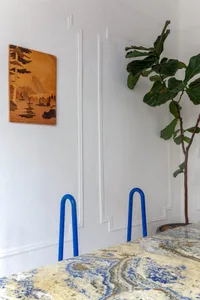 A modern interior featuring a marble table with blue-painted steel chairs, a fiddle leaf fig plant, and a small landscape pai