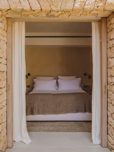 A minimalist bedroom with a bed covered in lavender linen sheets and a stone wall, enclosed by white curtains.