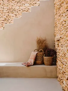 A minimalist interior featuring natural materials, including baskets and dried plants, against a stone wall and warm-toned wa