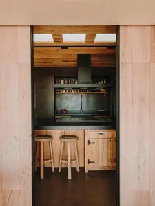 Modern kitchen with wooden ceiling and floor, featuring a black countertop, wooden stools, and a minimalist design.