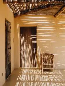 A sunlit room with woven chairs, wooden doors, and shadows cast by a woven lantern.