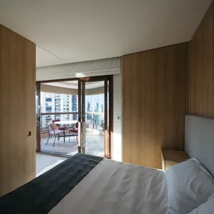 A modern minimalist bedroom featuring wooden walls and a white bed with gray accents, with a view of an outdoor seating area 