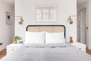 A minimalist white bedroom featuring a bed with a wicker headboard, white cotton bedding, and two wall-mounted brass lamps.
