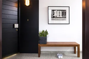 A modern entrance hall featuring a wooden bench, black doors, a wall-mounted light fixture, and a potted plant.