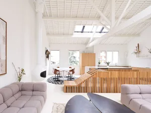 Modern living room with grey sofas, wooden flooring, and a white dining table surrounded by chairs. Natural light and white c