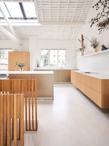 Modern kitchen features wooden cabinets and white flooring, with a minimalist design and large windows.