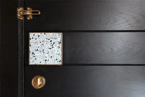 An elegant, black-colored wooden door with a geometric, white speckled terrazzo tile and a gold latch.