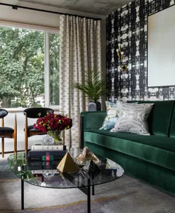 A modern living room featuring a green velvet sofa, glass coffee table, and patterned wallpaper.
