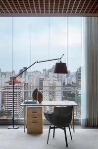 A minimalist office with a desk, chair, and floor lamp in front of a large window with a cityscape view.