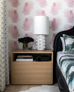 A minimalist bedroom featuring a black bedside table with a white lamp, black flowers, and books, placed against a wall with 