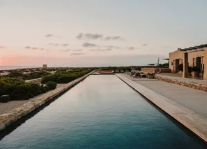 A minimalist vacation home featuring a long, rectangular swimming pool surrounded by a stone patio with outdoor furniture and