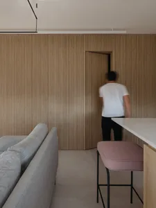 A modern residential kitchen featuring wood-paneled walls, a contemporary sofa, and a minimalist barstool with pink cushions.