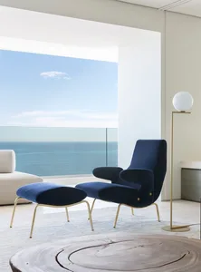 A minimalist living room with a blue velvet armchair, brass floor lamp, and a large window overlooking the ocean.