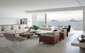 Modern living room with white and neutral-colored furniture and decor, featuring a large window overlooking the ocean.