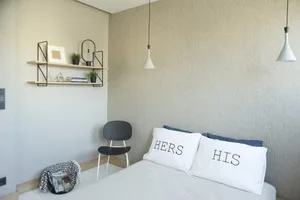 A minimalist bedroom with a bed, two pillows labeled 'HIS' and 'HERS', and a black chair with a shelf above it.