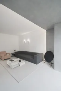 A minimalist living room with a concrete wall and floor, featuring a gray sofa, a white table, and a beige armchair.