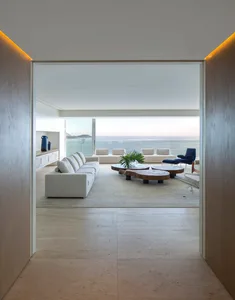 Modern white fabric sectional sofa and wooden coffee tables in minimalist living room overlooking the ocean.