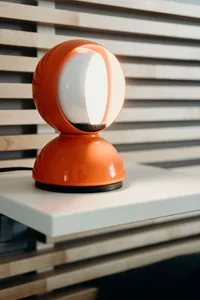 An orange table lamp with a white light bulb inside, placed on a white surface with light wood slats in the background.
