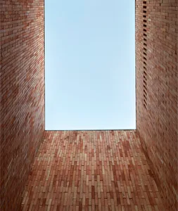 A rectangular room with brick walls and floor, looking up towards a blue sky through a window.