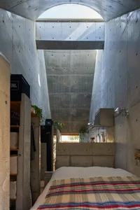 A minimalist bedroom featuring a concrete wall and floor, with a bed covered in a striped blanket and a skylight above.