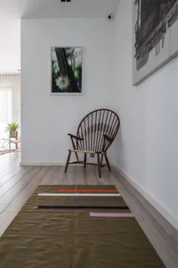A minimalist living room with a wooden floor, a single wooden chair, a colorful rug, and framed artwork on the walls.