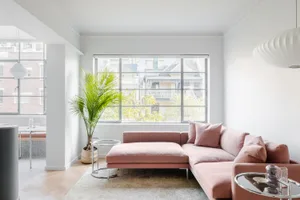 A modern, minimalist living room with a pink sofa, glass table, and large window overlooking urban buildings.