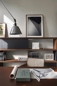 A modern office workspace featuring a wooden shelving unit with books, a sleek metal lamp, and various framed artworks.