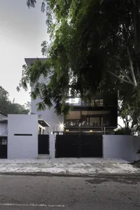 A modern residential building with a concrete and glass exterior, featuring a secure black gate and surrounded by lush greene