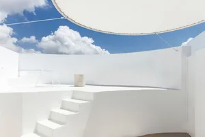 A minimalist white rooftop patio with concrete stairs and a white wire chair, under a circular awning and a blue sky with clo