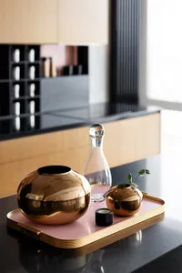 A modern kitchen counter featuring a pink tray with gold-trimmed bronze vases, a glass carafe, and a candle holder.