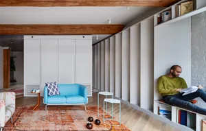A minimalist living room with a blue sofa, side table, and rug, featuring a bookshelf with books and a man sitting and readin