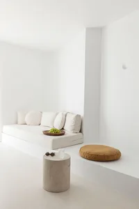A minimalist living area featuring a white couch against a white wall, a concrete cylinder table, and a brown cushion.