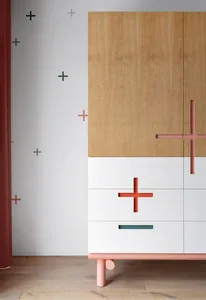 Modern minimalist wardrobe cabinet with wooden and white panels, orange and green crosses, and pink legs.