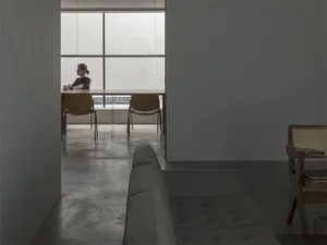 A minimalist modern coffee shop with concrete flooring, a long table, and a woman drinking coffee by a large window.