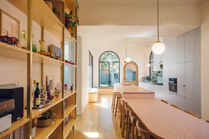 A modern kitchen featuring a pink terrazzo countertop and stools next to a wooden table and bookshelves.
