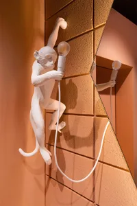 A minimalist wall sculpture of a white monkey holding a light bulb on a textured gold background.
