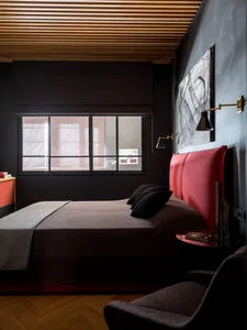 A modern minimalist bedroom featuring a red upholstered bed, dark walls, and a wooden ceiling.