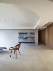 A modern living room with concrete flooring, a wooden chair, and a concrete wall with shelves.
