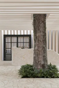 A modern entrance porch featuring a large tree surrounded by plants on a concrete patio with a stone wall and black-framed gl