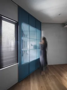 A modern minimalist bedroom featuring a large window with closed blinds, a blue perforated metal panel wall, and a blurred hu