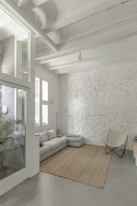 Minimalist living room with concrete walls and floor, featuring a white couch and a woven rug.