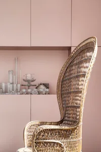 Modern wicker armchair with high backrest in a room with pale pink furniture, showing a collection of decorative glassware on