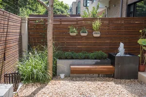 A modern urban garden featuring a wooden fence, concrete seating area, and pebble pathway with a water fountain.